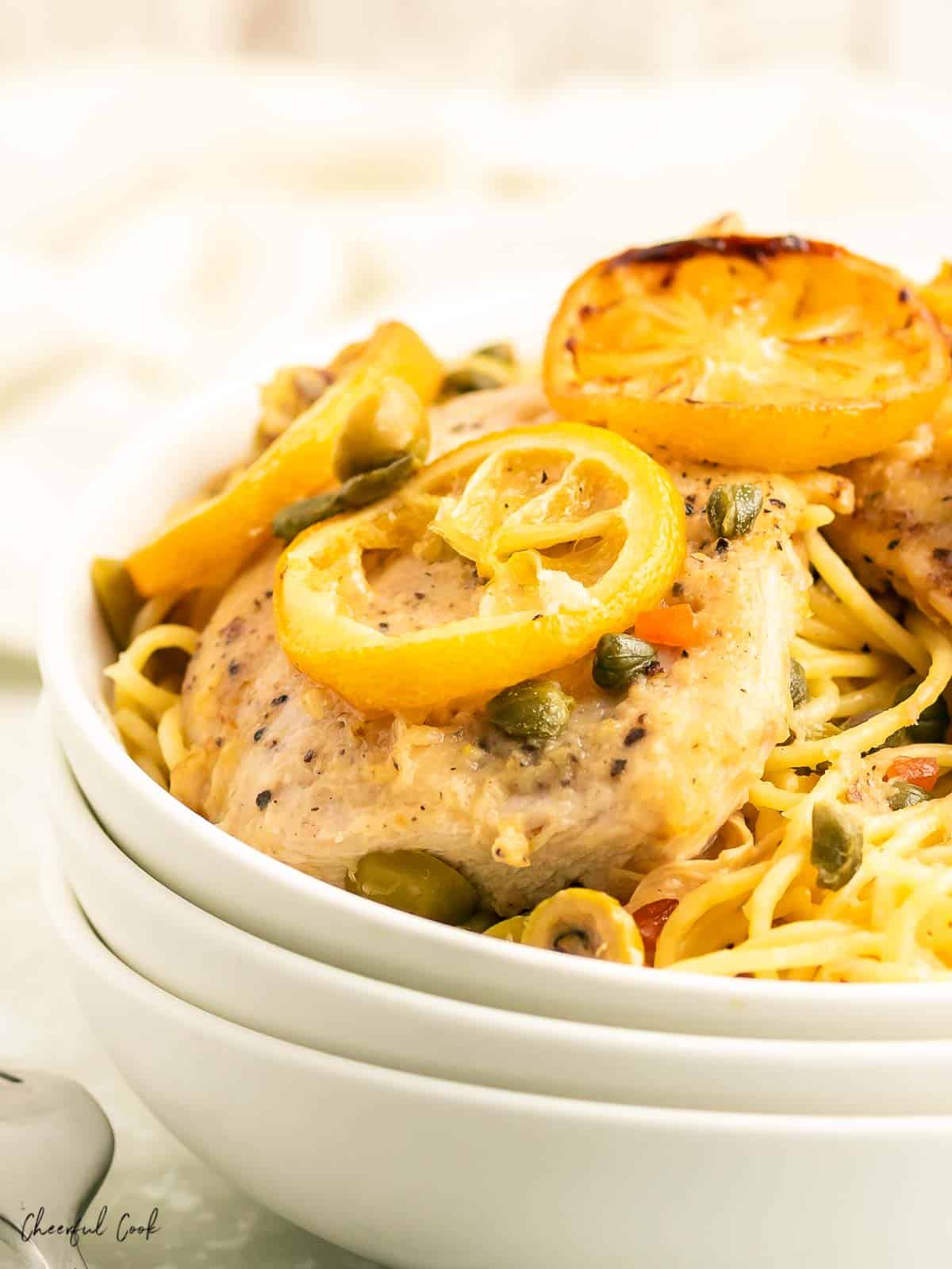Lemon Chicken in a caper, olive butter sauce topped over spaghetti pasta in a white bowl