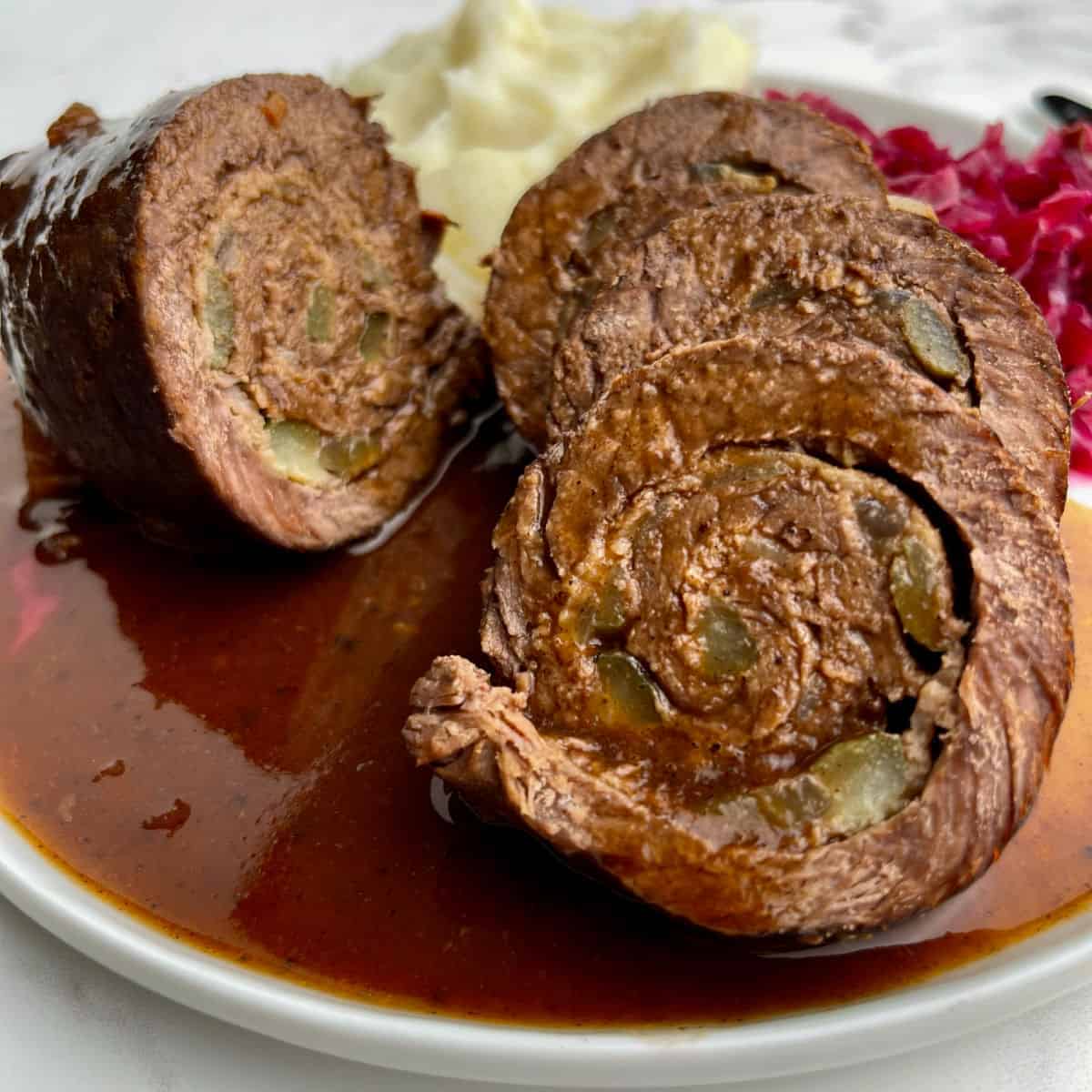 sliced freshly cooked German rouladen on a white plate with mashed potatoes and red cabbage