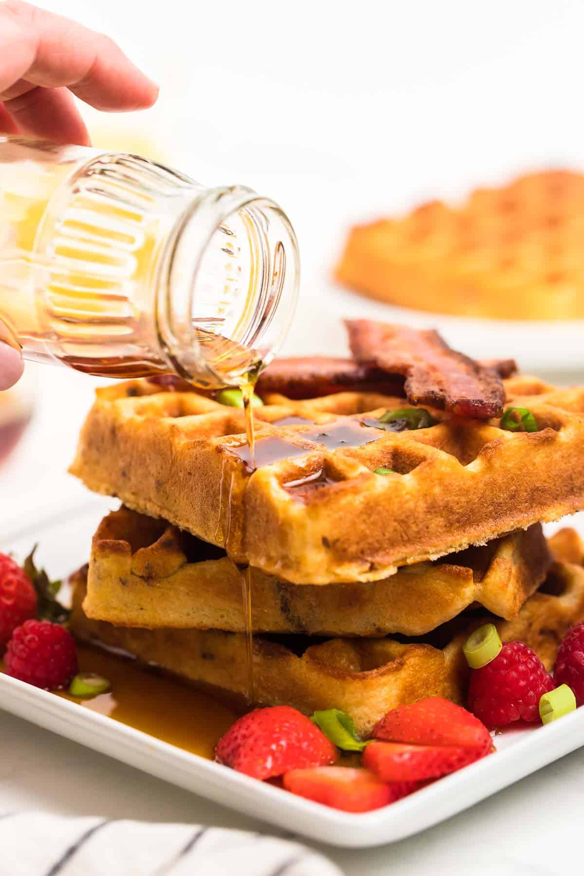 Drizzling maple syrup over cornmeal waffles with bacon sitting on a white plate
