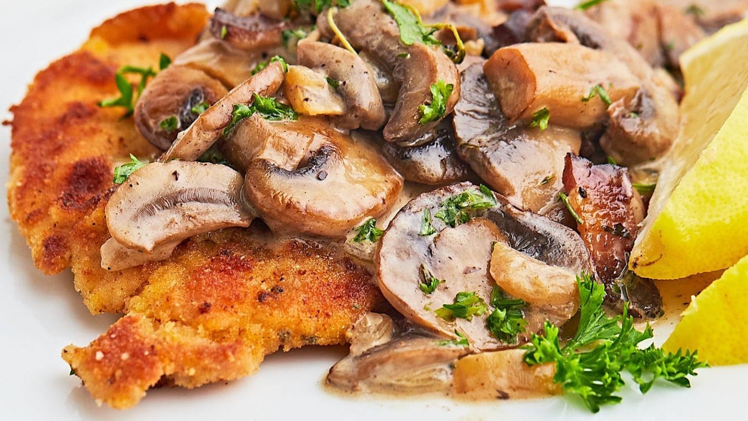 A breaded chicken cutlet topped with saut&eacute;ed mushrooms and herbs, served with lemon wedges on a white plate.