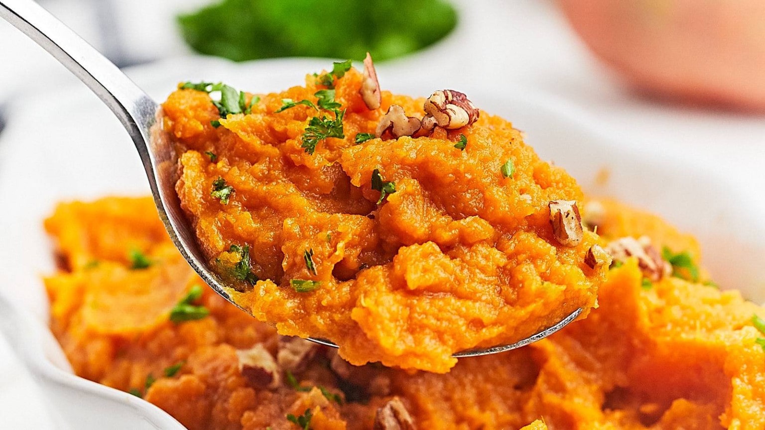 A spoonful of creamy sweet potato mash topped with chopped pecans and parsley, set against a blurred green background.