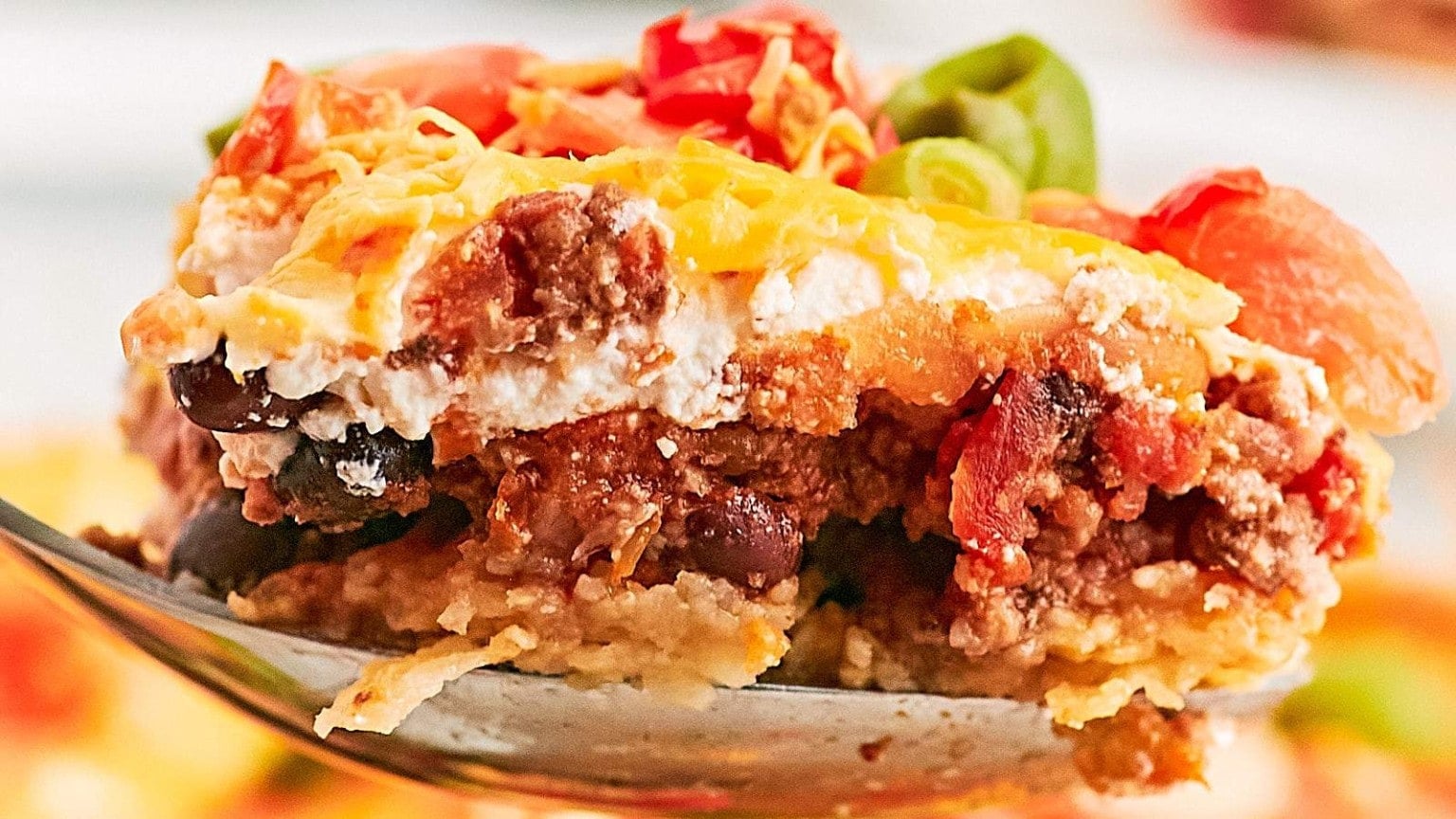 A close-up of a layered casserole scoop featuring ground beef, black beans, cheese, tomatoes, and green onions.