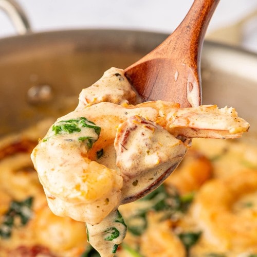 A wooden spoon is being used to scoop shrimp and spinach out of a pan.