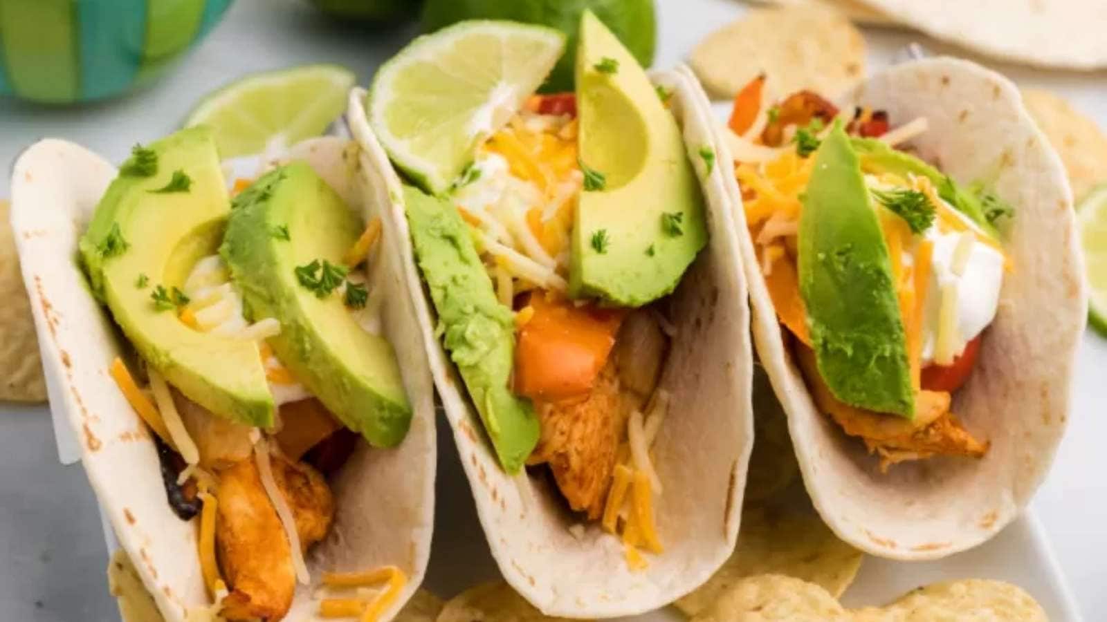 Three chicken tacos with avocado and lime on a white plate.