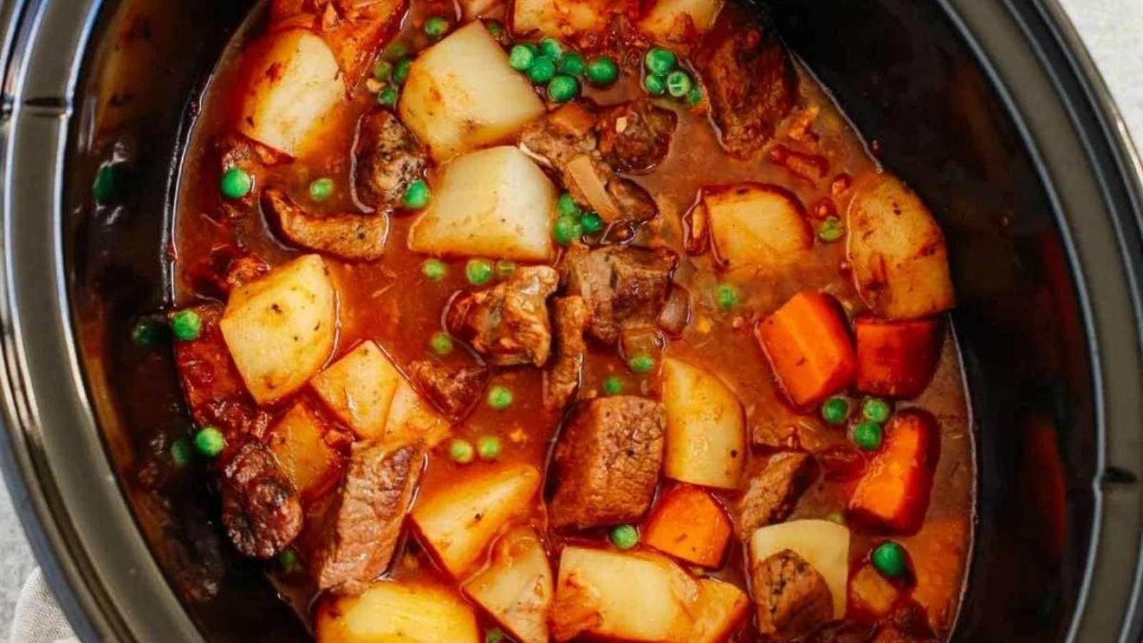 A crock pot full of beef, potatoes and peas.