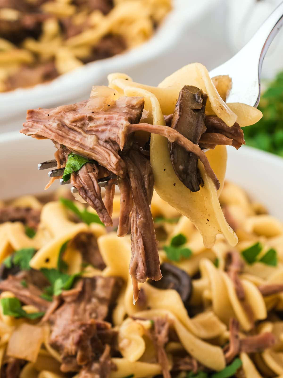 A fork is being held up to a plate of pasta with beef and mushrooms.