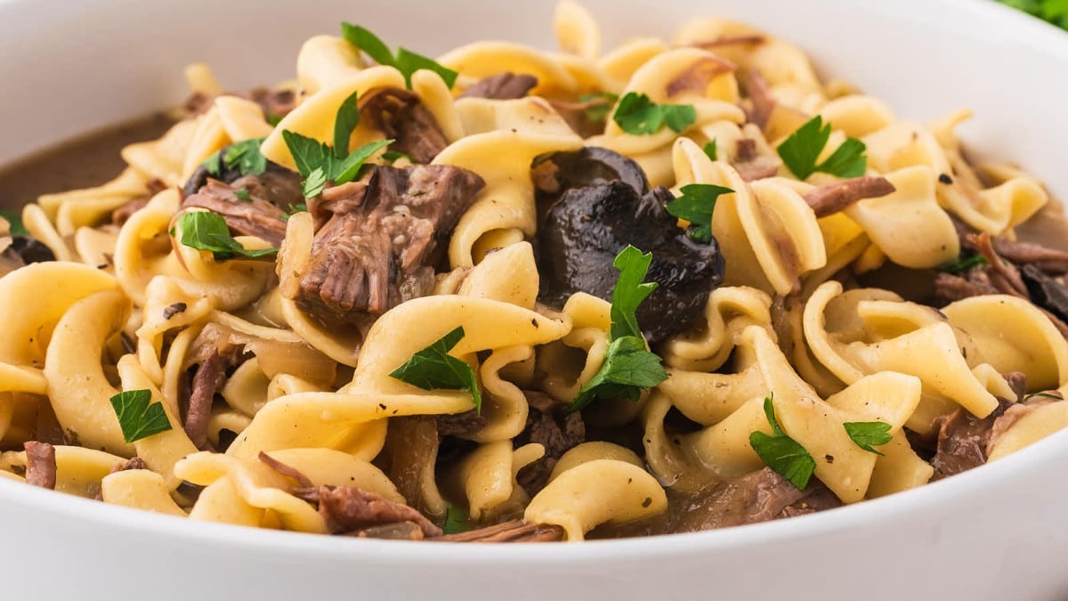 A close-up of a bowl of beef stroganoff featuring tender egg noodles, chunks of beef, mushrooms, and a creamy sauce, garnished with fresh parsley. The dish appears rich and savory, with a comforting homemade appeal.