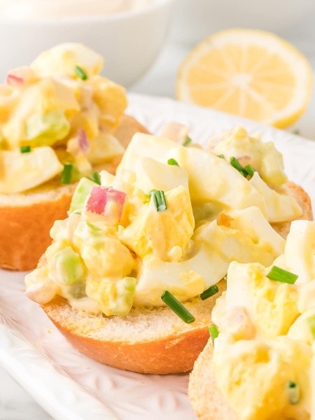 Egg salad sandwich on a white plate with lemon wedges.