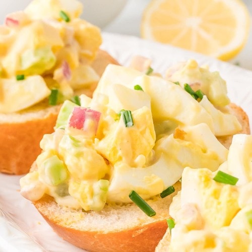 Egg salad sandwich on a white plate with lemon wedges.