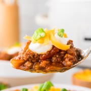A spoonful of chili with cheese and sour cream in front of a slow cooker.