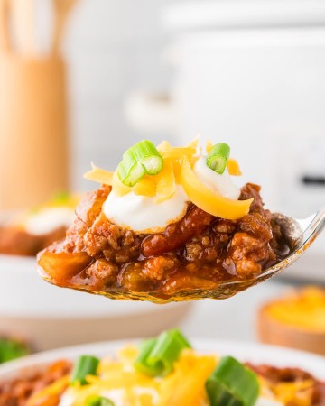 A spoonful of chili with cheese and sour cream in front of a slow cooker.