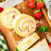 A strawberry roll with whipped cream and strawberries on a cutting board.