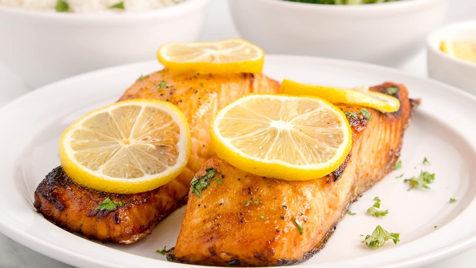Two grilled salmon fillets garnished with fresh lemon slices, served on a white plate with herbs, alongside bowls of rice and greens.