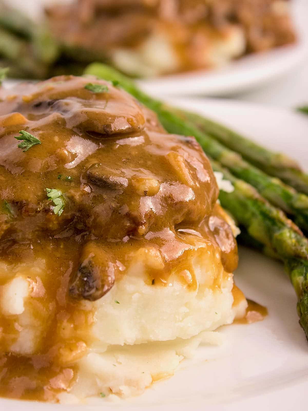 Slow Cooker Salisbury Steak served over mashed potatoes with green asparagus on a white plate.