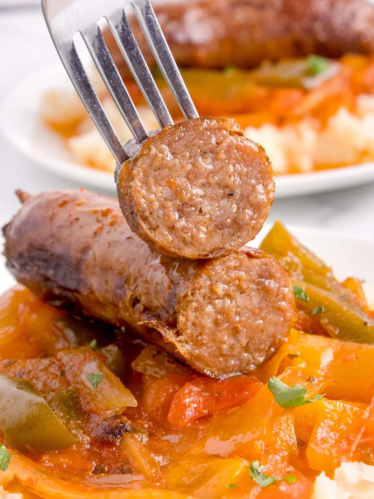A fork holding a sliced piece of sausage that is sitting on cooked peppers.