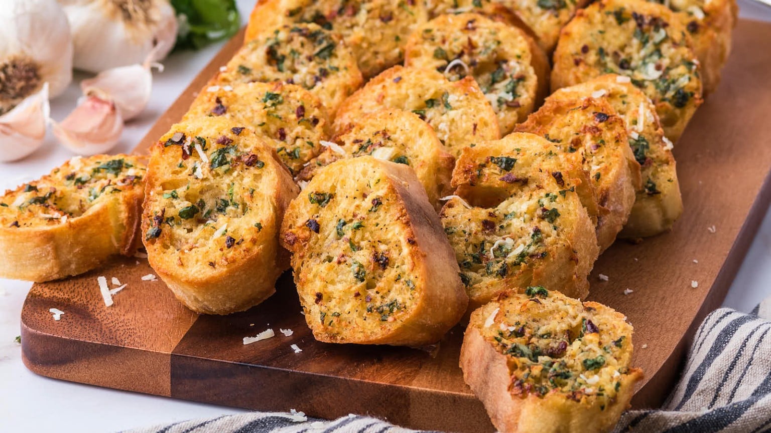 Garlic bread slices topped with herbs and cheese, arranged on a wooden board with garlic cloves in the background.