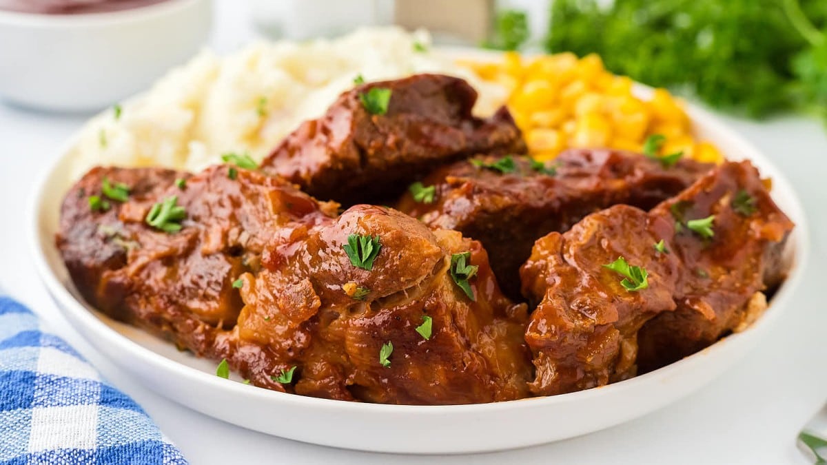 A plate of tender, saucy ribs garnished with herbs, accompanied by creamy mashed potatoes and bright yellow corn. A checkered napkin is nearby, and fresh greens are blurred in the background.