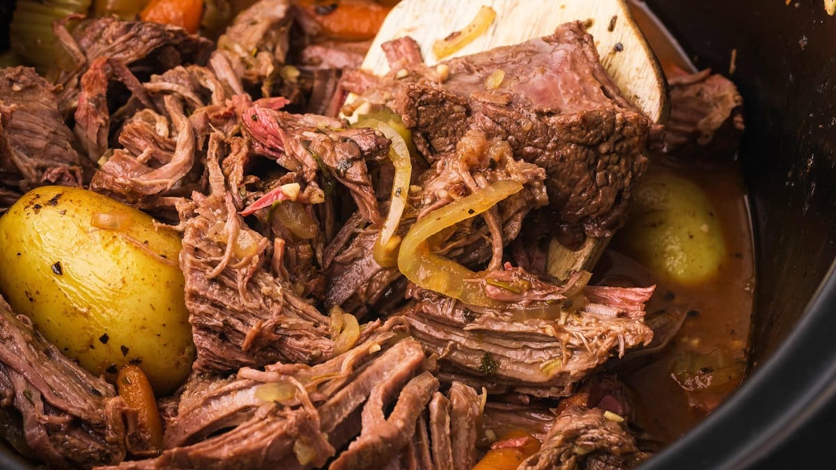 A close-up view of a pot roast with shredded beef, onions, carrots, and potatoes. The tender meat is mixed with vegetables and is being stirred with a wooden spoon. The dish is rich in texture and looks flavorful.