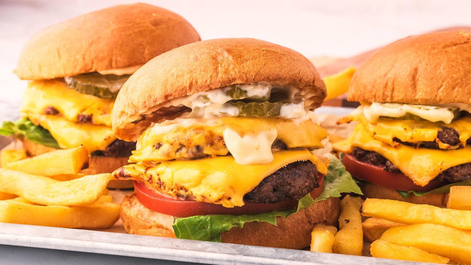 Three juicy cheeseburgers layered with lettuce, tomato, pickles, and creamy sauce, served with golden fries on a metal tray.