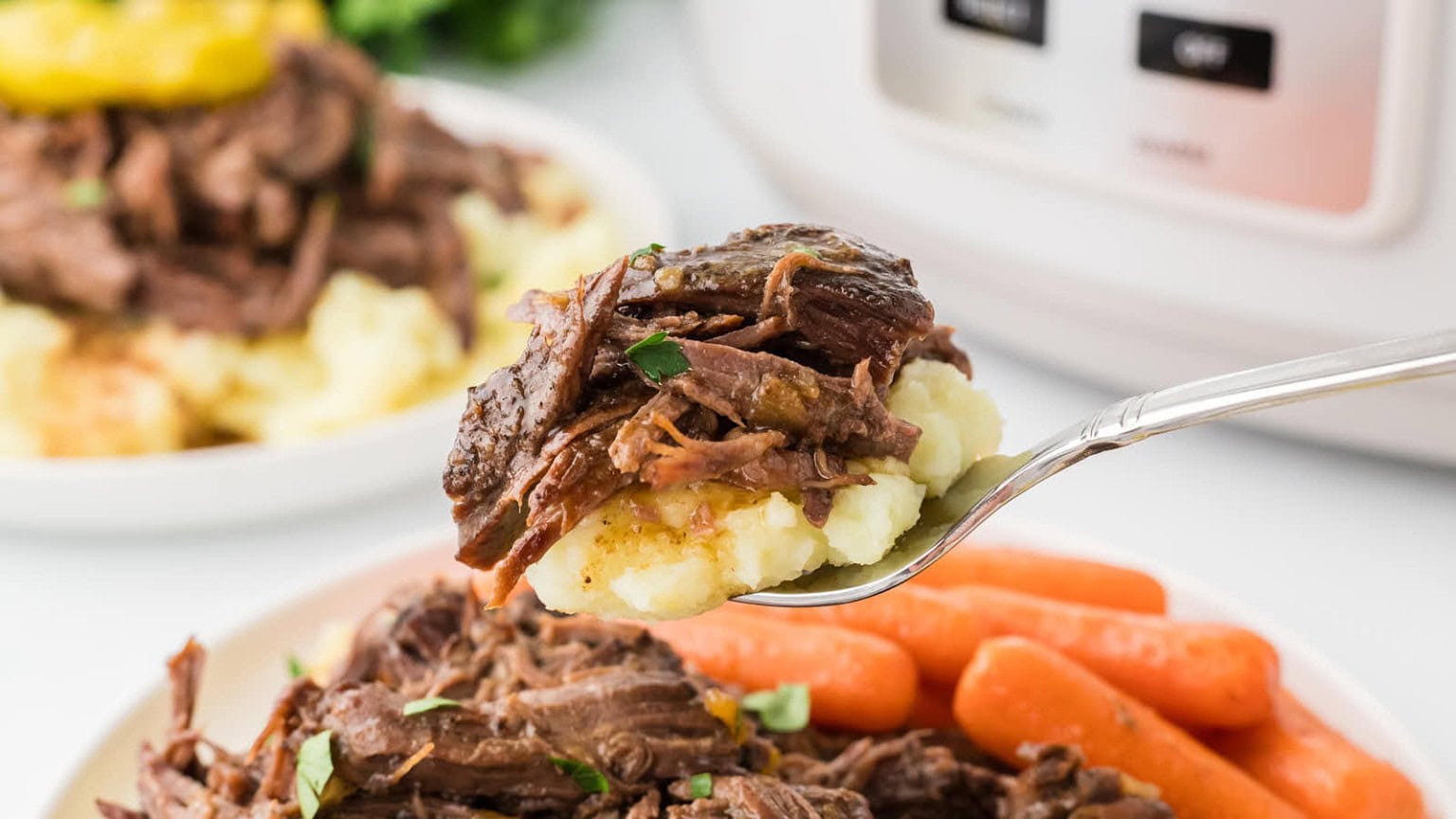 A fork holds tender beef over creamy mashed potatoes, with carrots in the foreground and a blurred plate in the background.