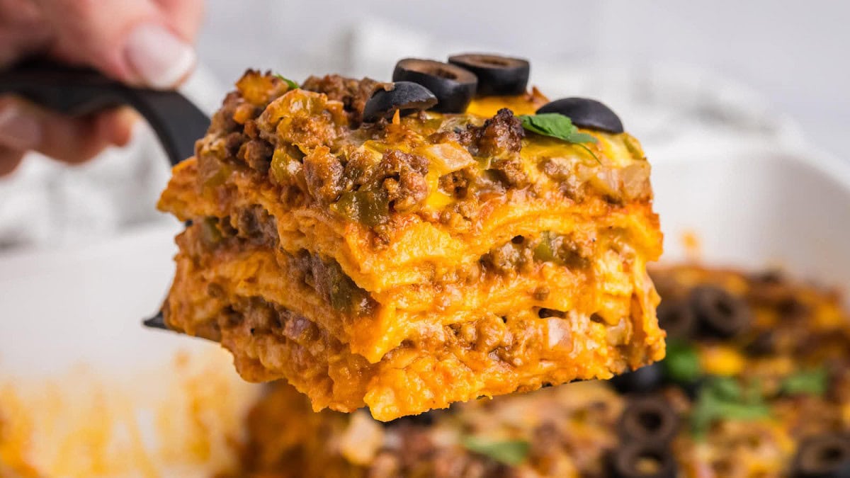 A spatula lifts a hearty slice of lasagna from a baking dish. The lasagna is layered with melted cheese, seasoned ground beef, and vegetables. Slices of black olives and fresh basil leaves garnish the top layer. The background shows the remaining lasagna in the dish.