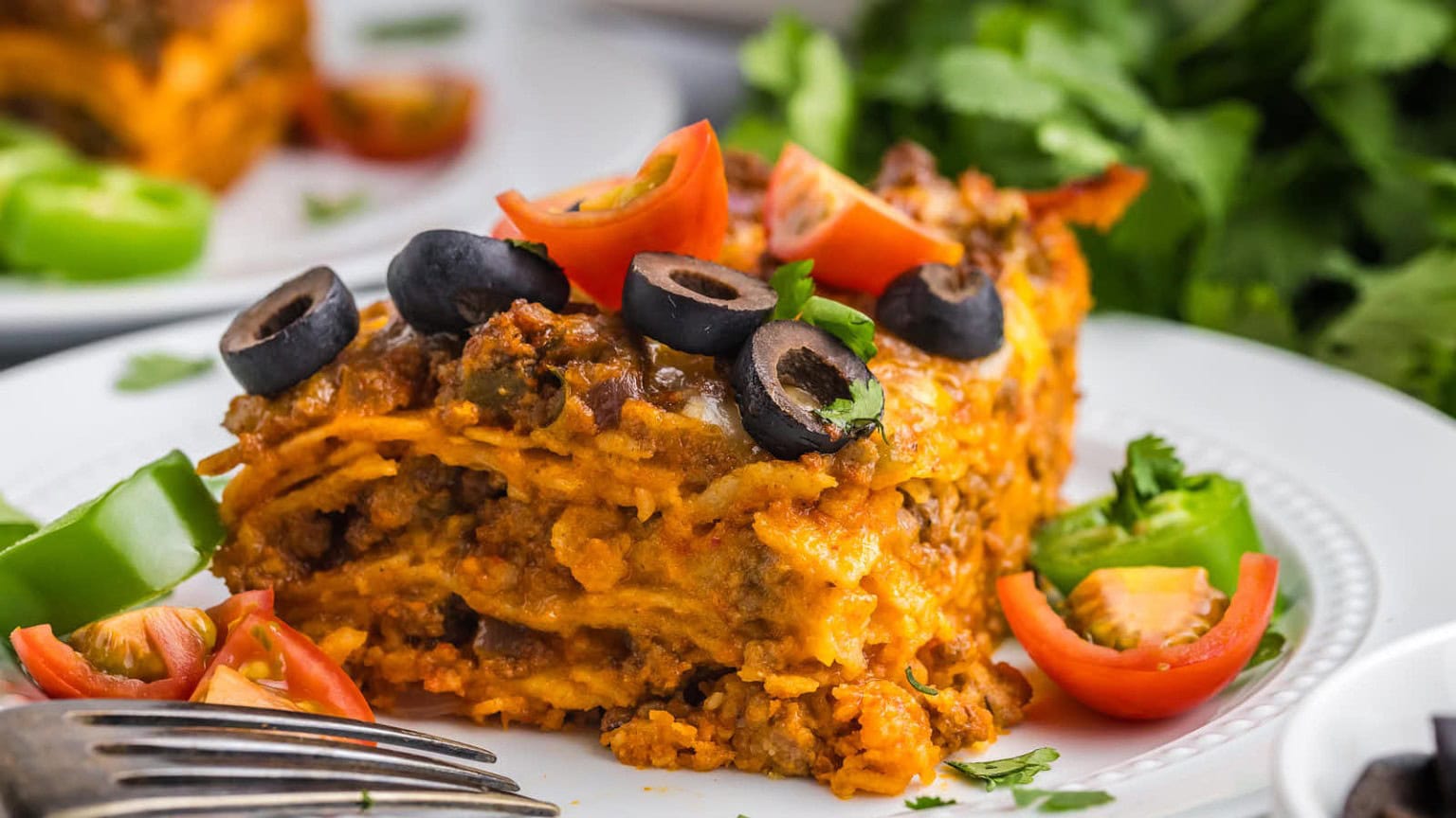 A delicious slice of layered lasagna topped with olives, tomatoes, and fresh cilantro, served with green bell pepper and cherry tomatoes.