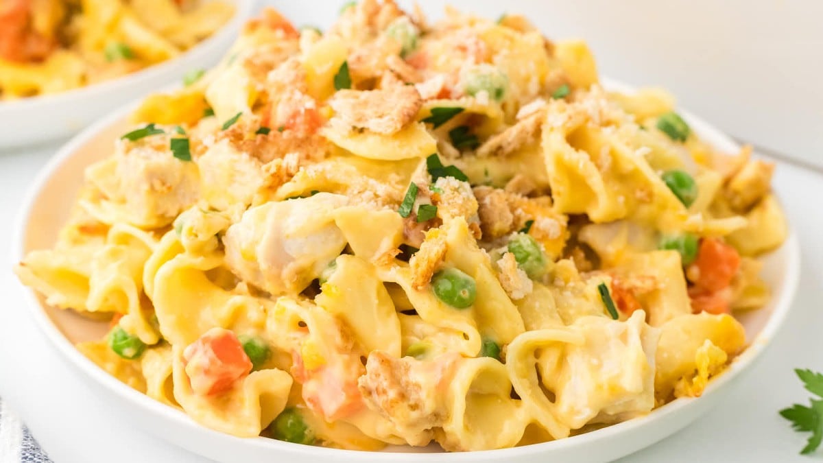 A close-up of a plate of creamy chicken noodle casserole, featuring broad egg noodles, chunks of chicken, peas, and diced carrots, all topped with a golden breadcrumb crust and garnished with chopped parsley.