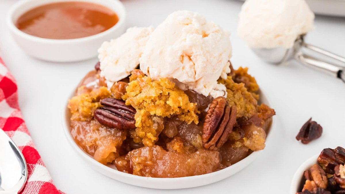 A dessert plate with warm apple cobbler topped with golden brown crumble, crunchy pecans, and scoops of vanilla ice cream. A small bowl of caramel sauce and an ice cream scoop are in the background.