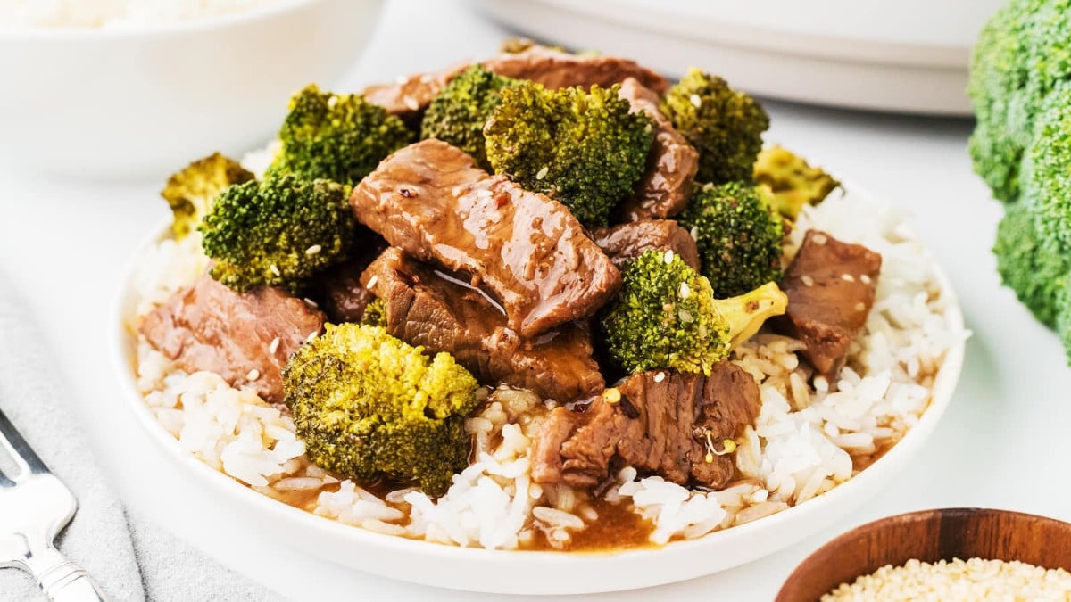 A plate of beef and broccoli stir-fry served over a bed of white rice. The beef is glazed with a rich, brown sauce and is mixed with vibrant green broccoli florets. A fork and a small bowl of sesame seeds are nearby.
