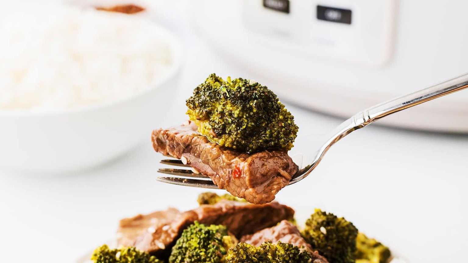 A fork lifts a piece of tender beef topped with broccoli, with a bowl of white rice blurred in the background.