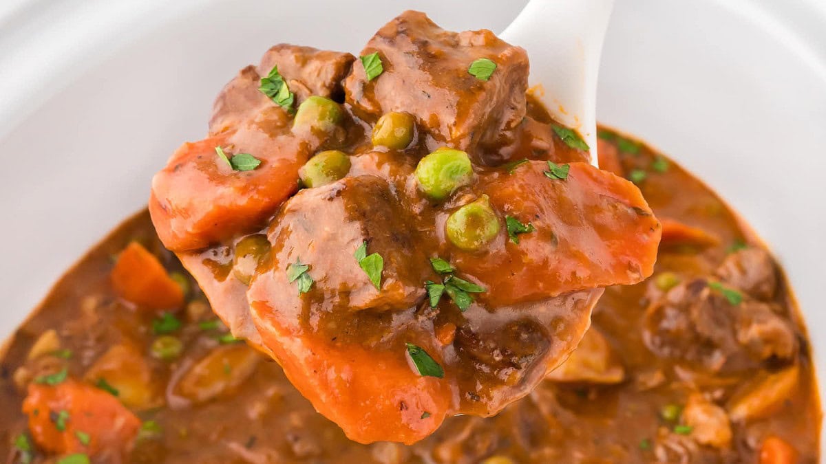 A spoonful of beef stew with chunks of beef, carrots, peas, and garnished with fresh herbs. The stew has a rich, brown sauce.
