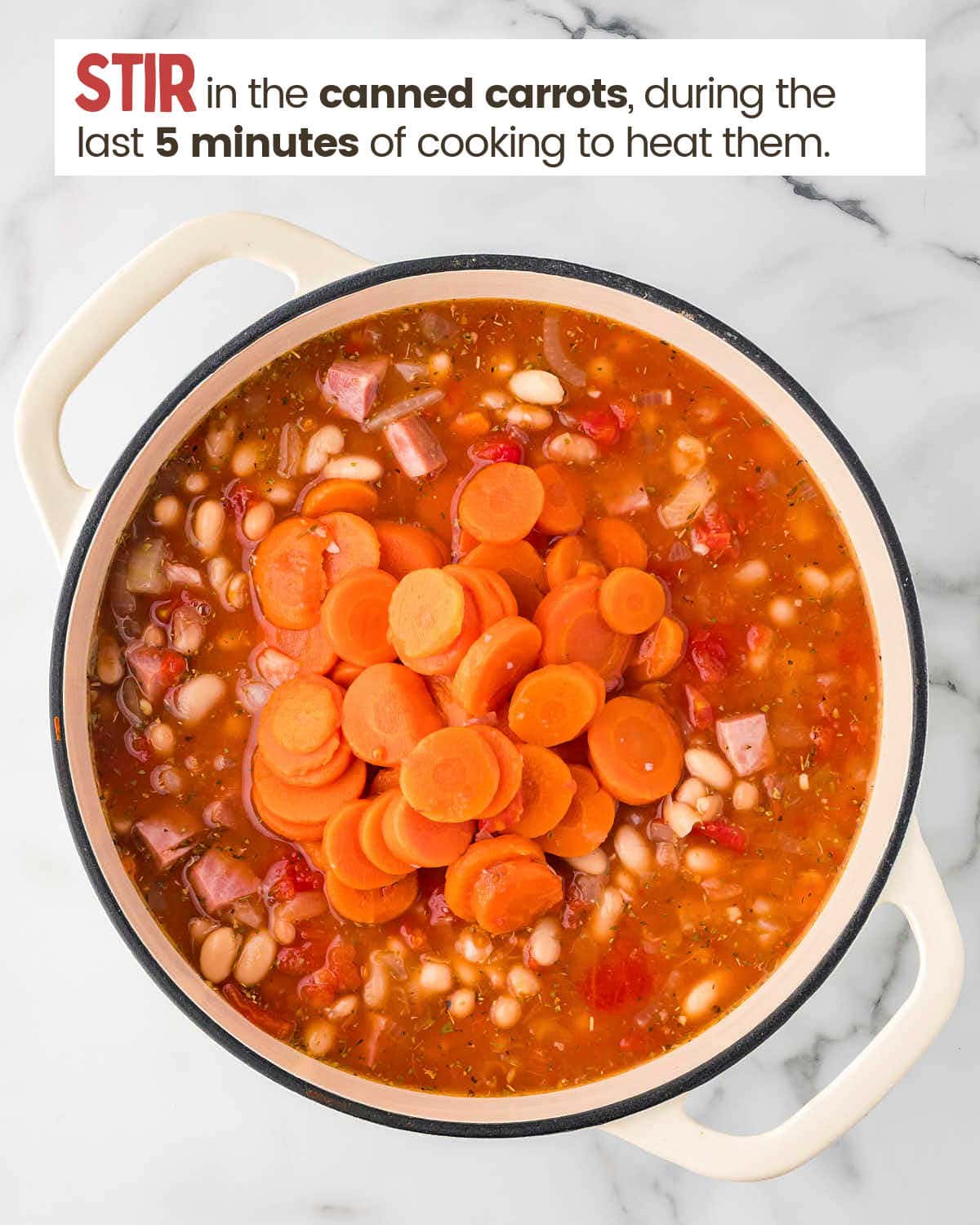 Adding canned carrots to ham and bean soup in a large pot.