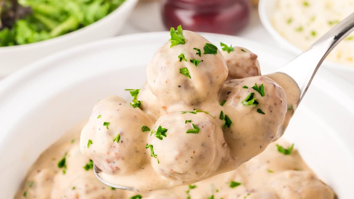 A spoonful of creamy, herb-garnished meatballs over a bowl filled with similar meatballs. In the background, there are blurred elements suggesting a salad and possibly a sauce bottle.