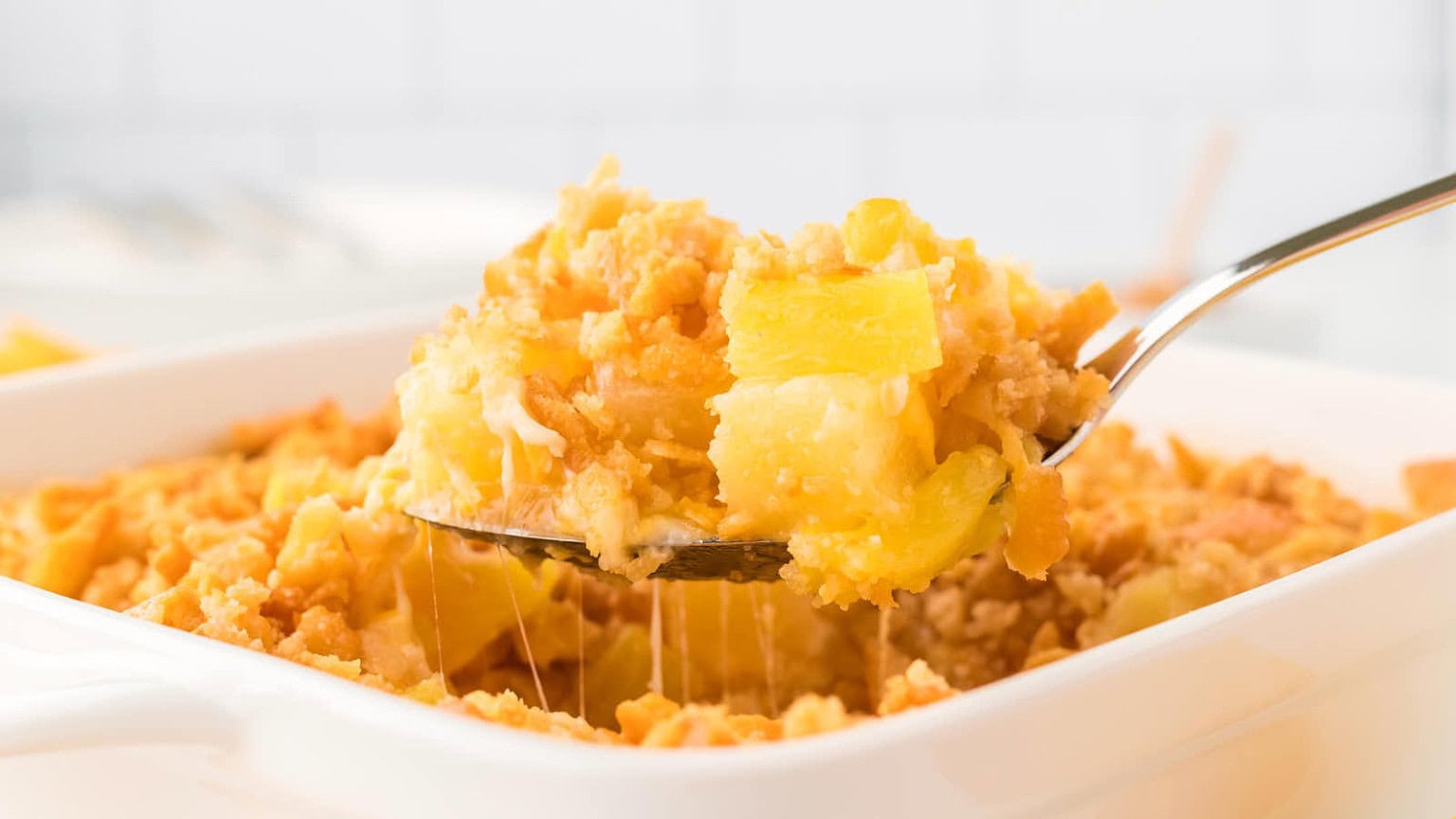 A silver spoon lifting a cheesy, golden-brown casserole with pineapple chunks from a white baking dish.