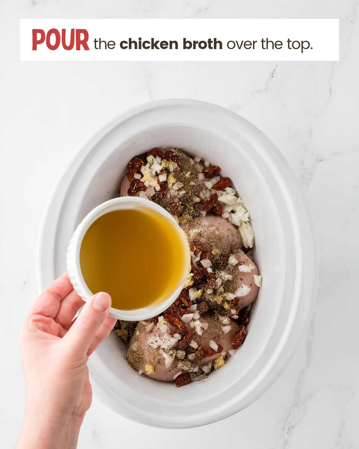 A hand holding a dish with broth in it over chicken breasts with garlic and spices in a slow cooker.