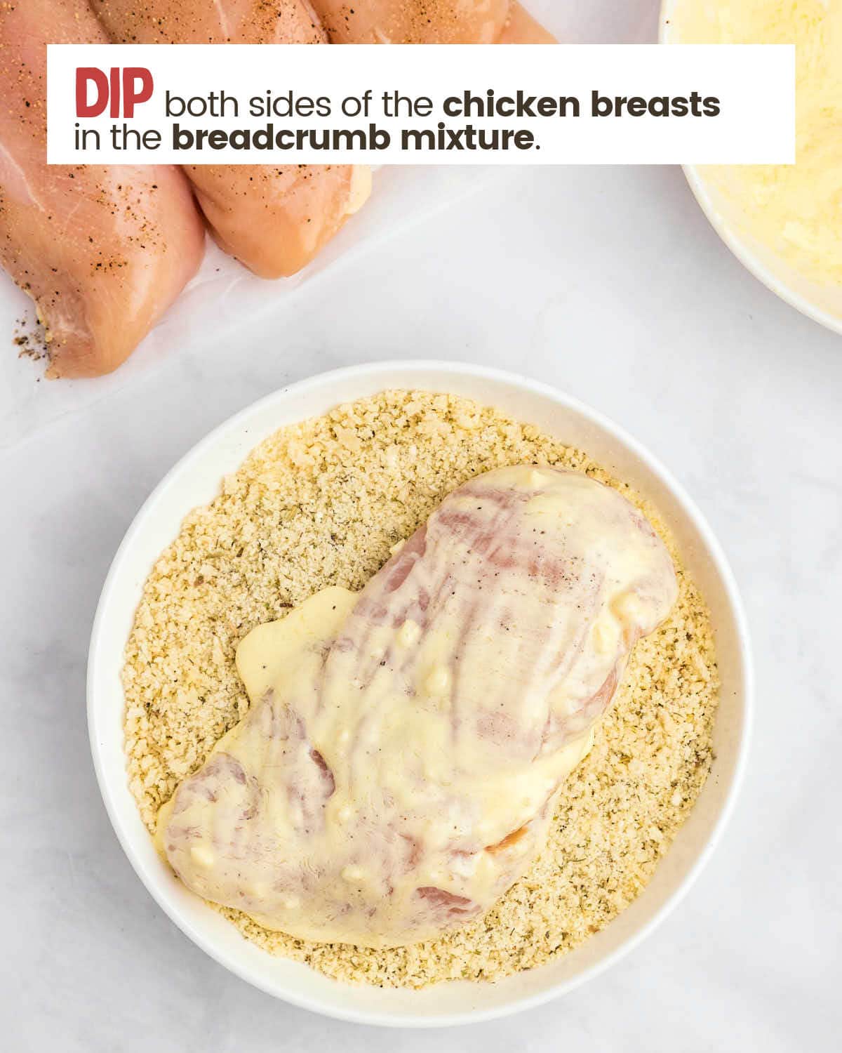 A chicken breast coated with a mayo/butter mixture is in a bowl of seasoned breadcrumbs.