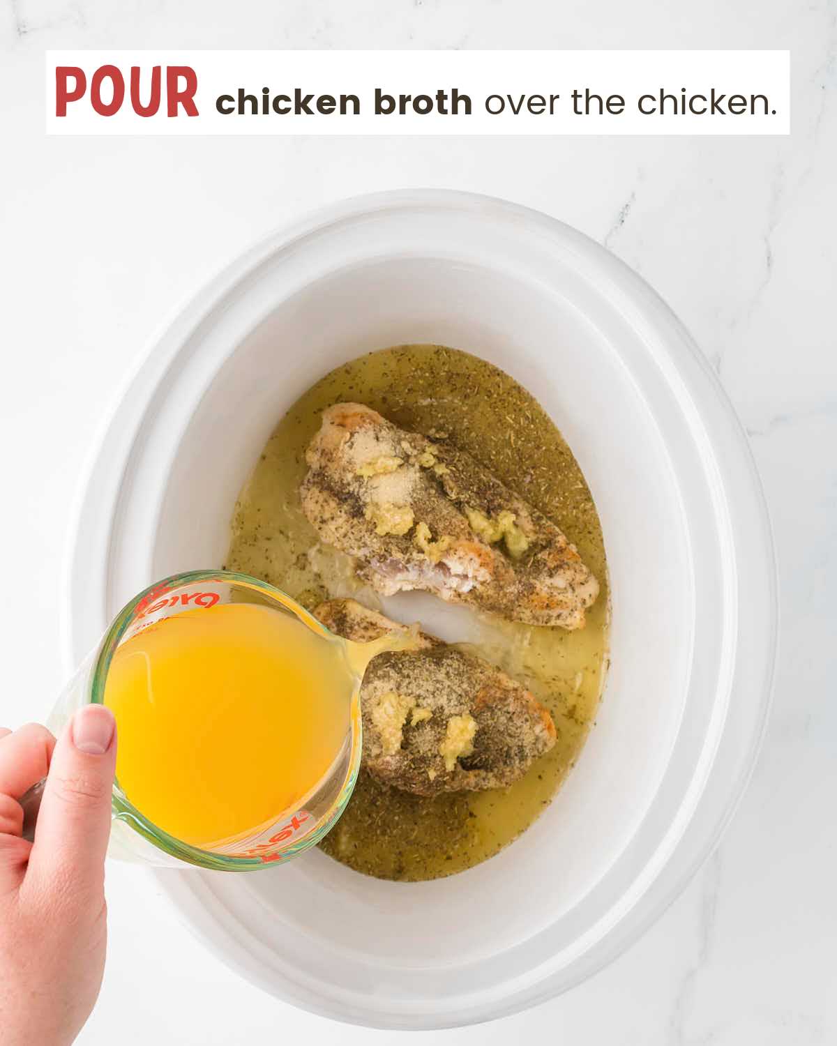 A hand pouring chicken broth from a measuring cup over chicken in a slow cooker.