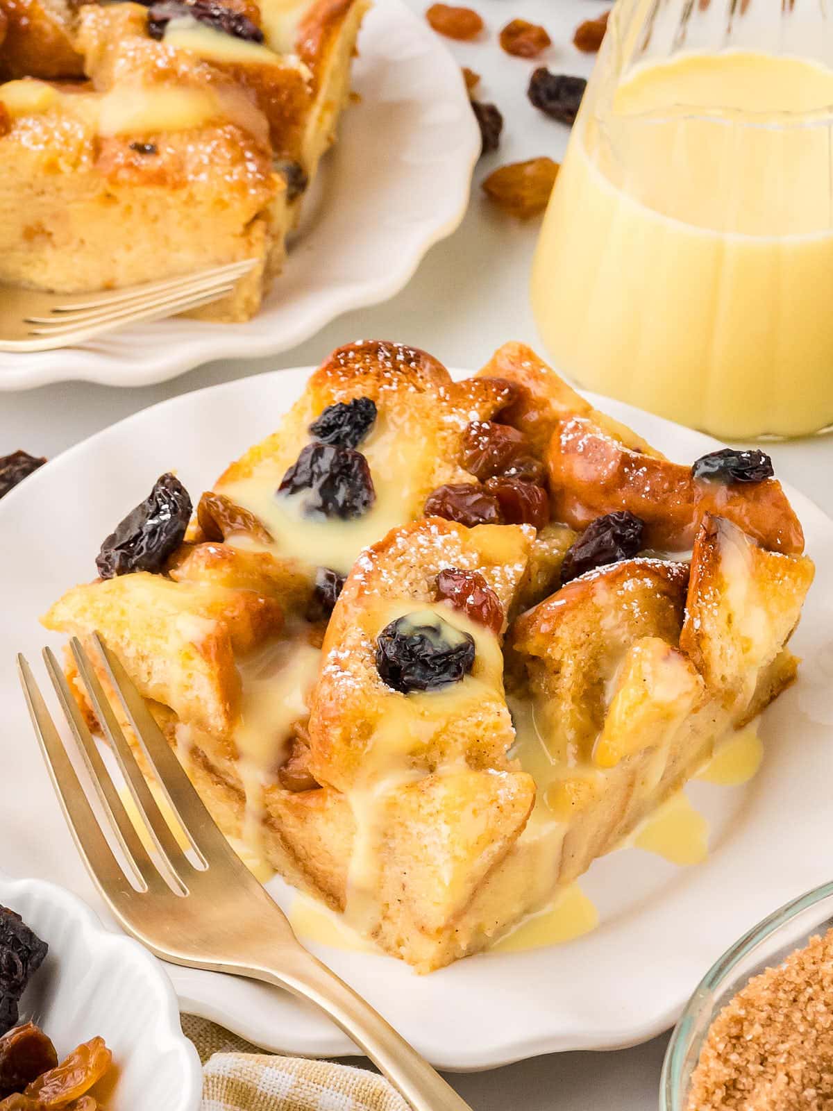 A slice of Bread Pudding on a white plate drizzled with warm vanilla sauce.
