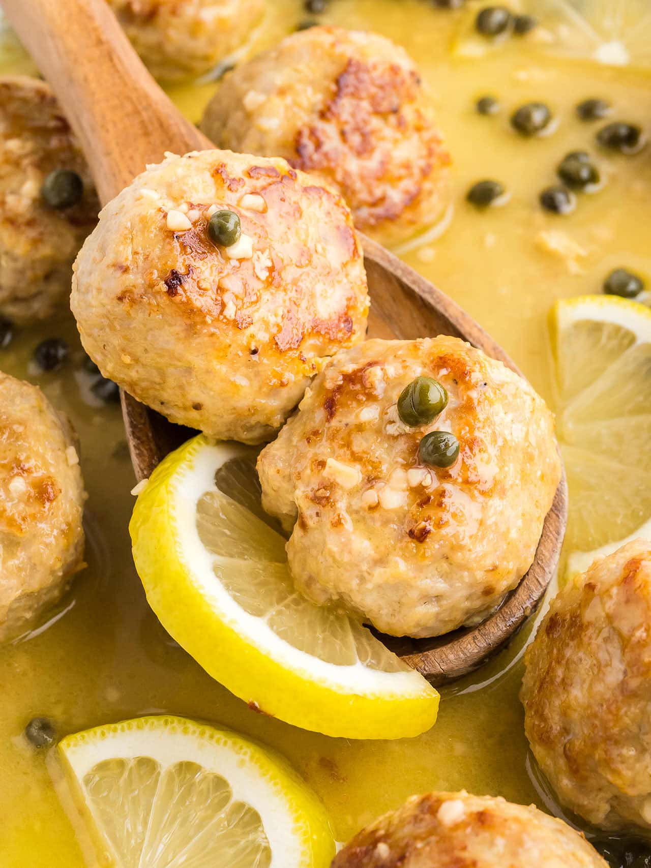 A wooden serving spoon lifting two Chicken Piccata Meatballs out of the skillet.