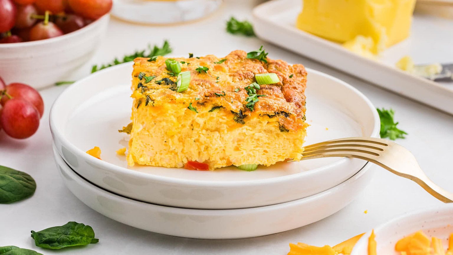 A slice of egg frittata topped with green onions, on stacked white plates, surrounded by spinach and grapes.