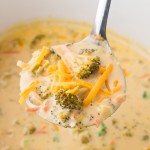 A ladle of Cheddar Broccoli Soup in a white Crockpot.