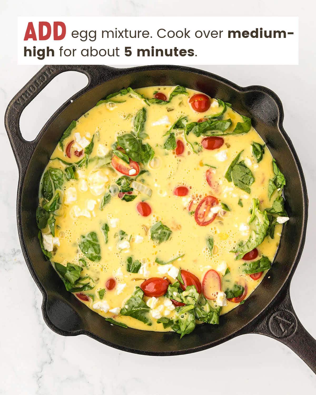 Eggs mixed with feta cheese, spinach, and cherry tomatoes in a cast iron pan.