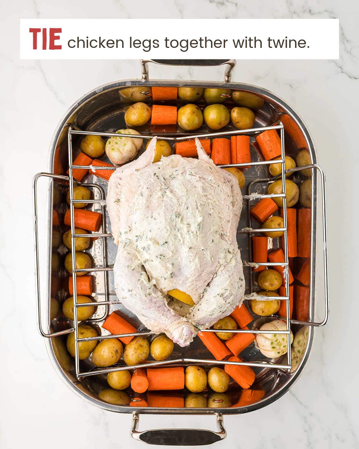 A butter mixture on a chicken with its legs tied with twine on a roasting rack over potatoes and carrots.