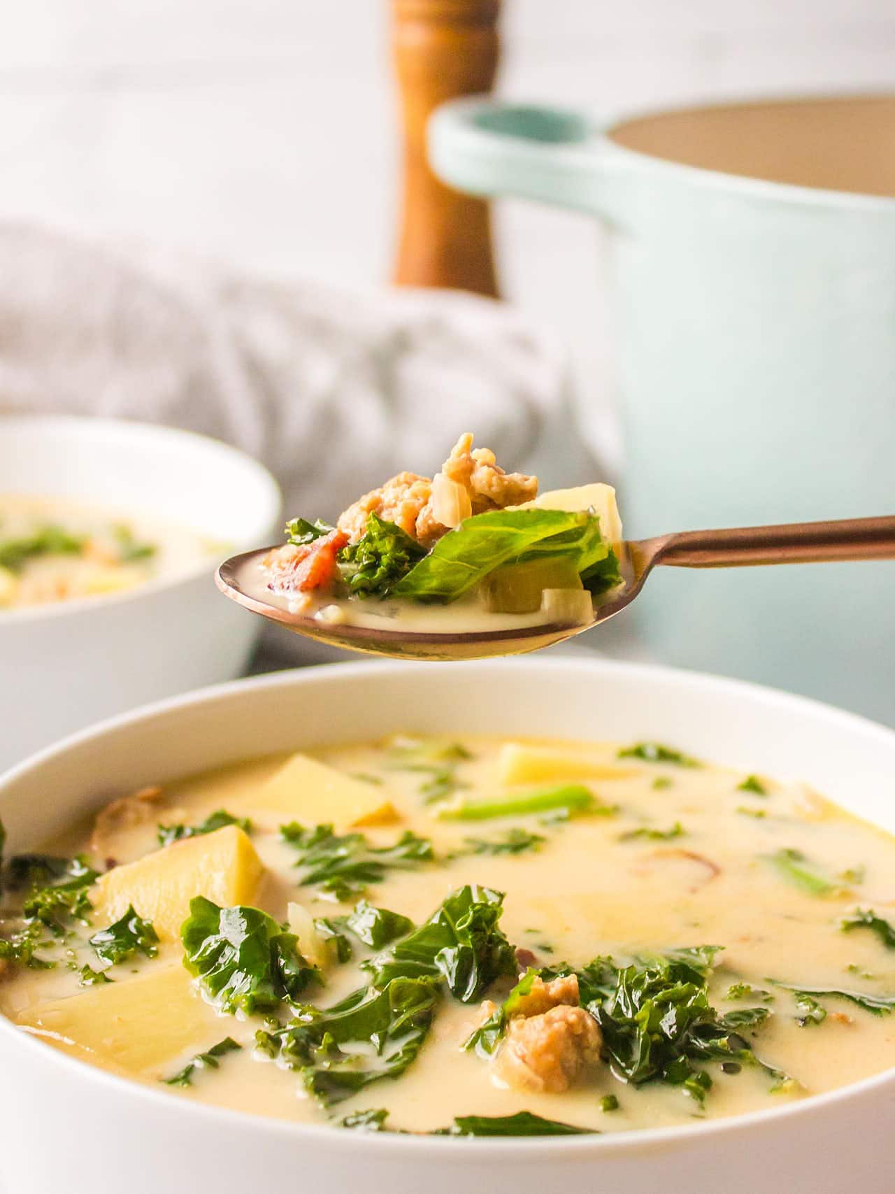 A spoonful of Zuppa Toscana hovering over a white serving bowl.