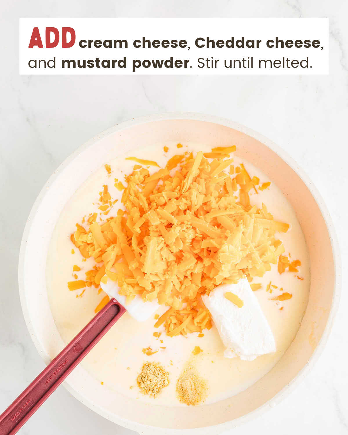 Shredded cheese, cream cheese, and mustard powder over a spatula in a cream mixture in a skillet.