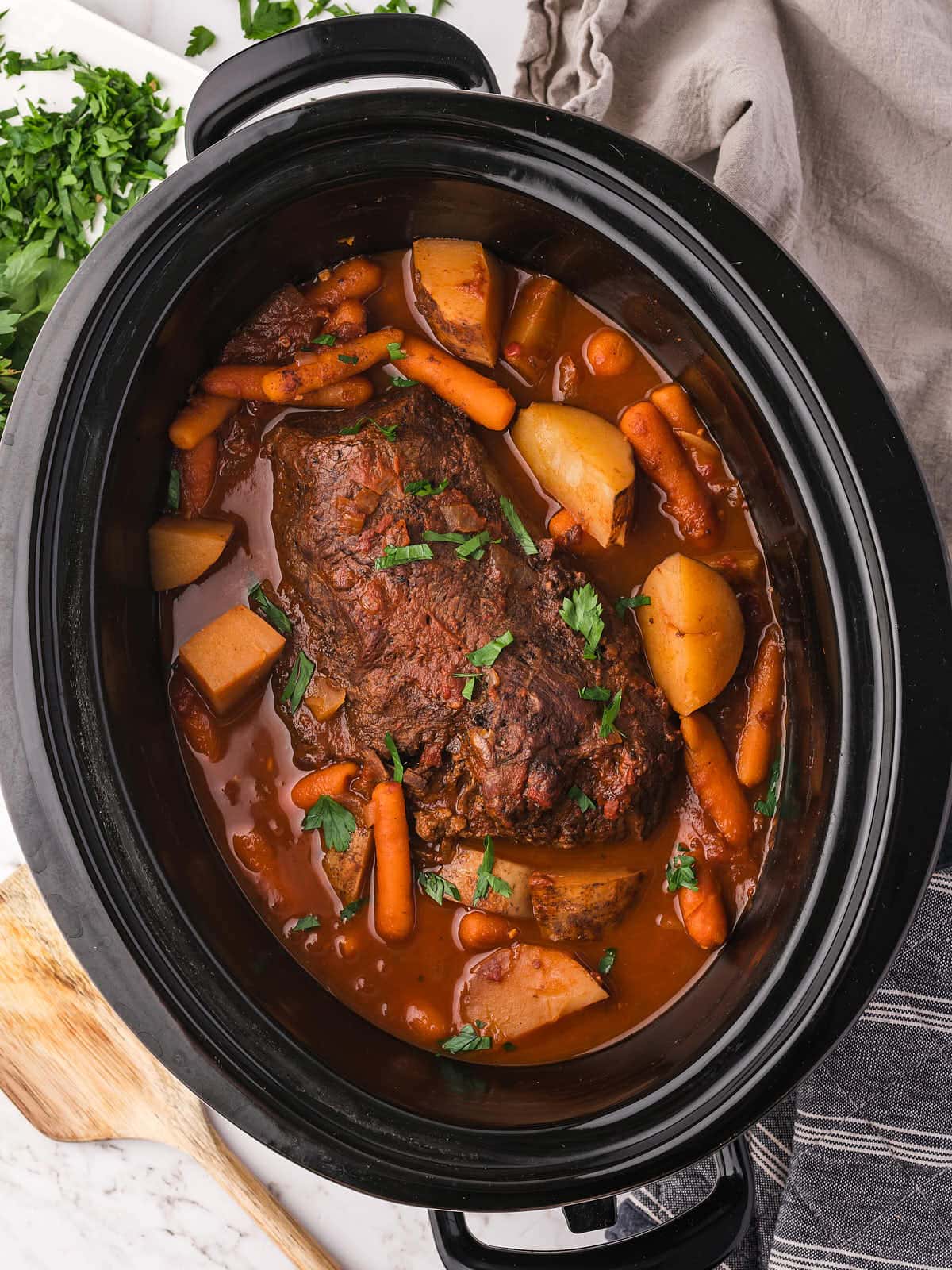 Slow cooker pot roast with baby carrots and potatoes in a red wine tomato sauce topped with fresh parsley.