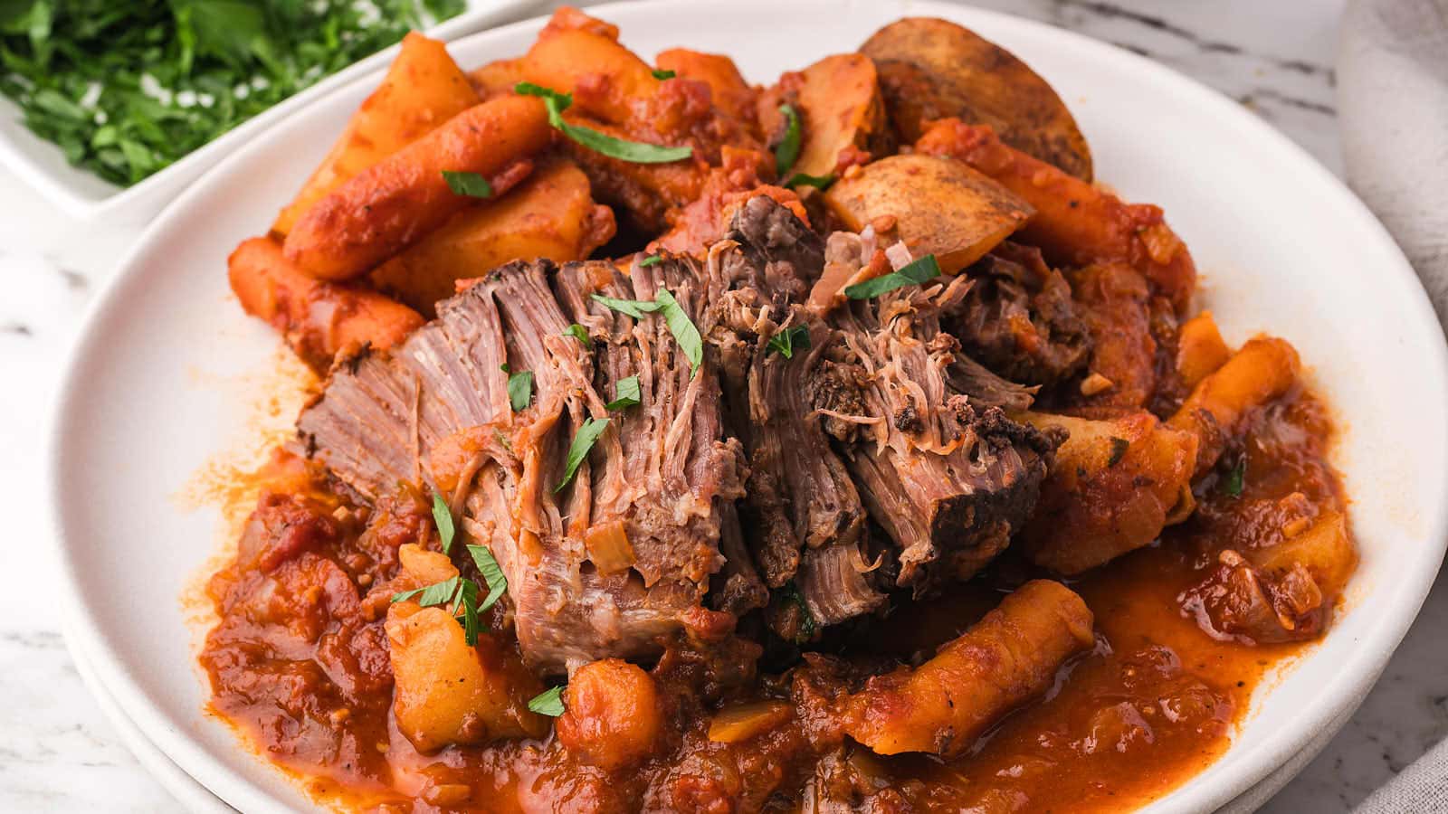 Fork-tender slow cooker pot roast pulled apart on a plate with carrots, potatoes, and tomato sauce.
