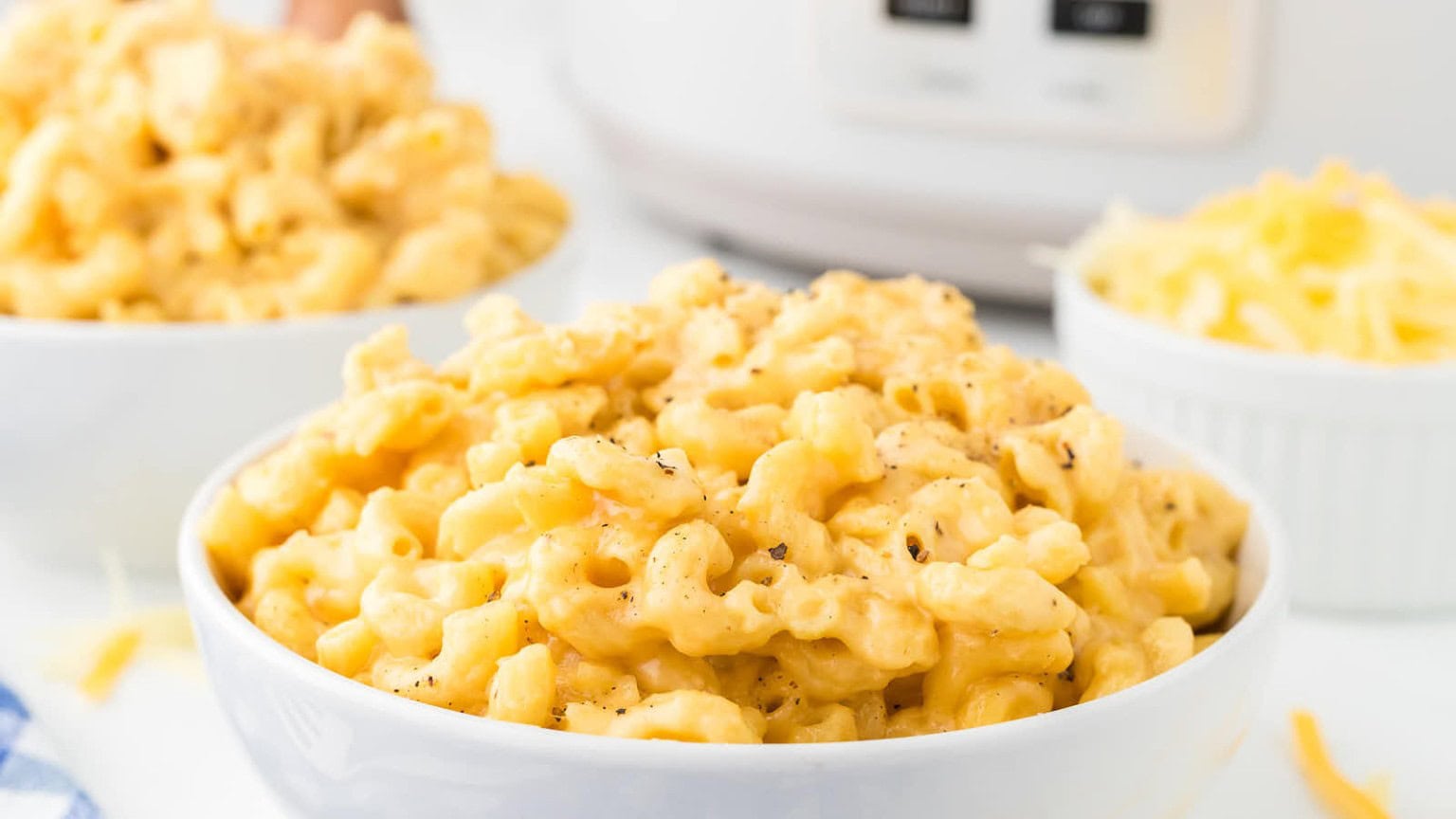 A close-up of creamy mac and cheese in a white bowl, with two bowls of macaroni and cheese and shredded cheese in the background.
