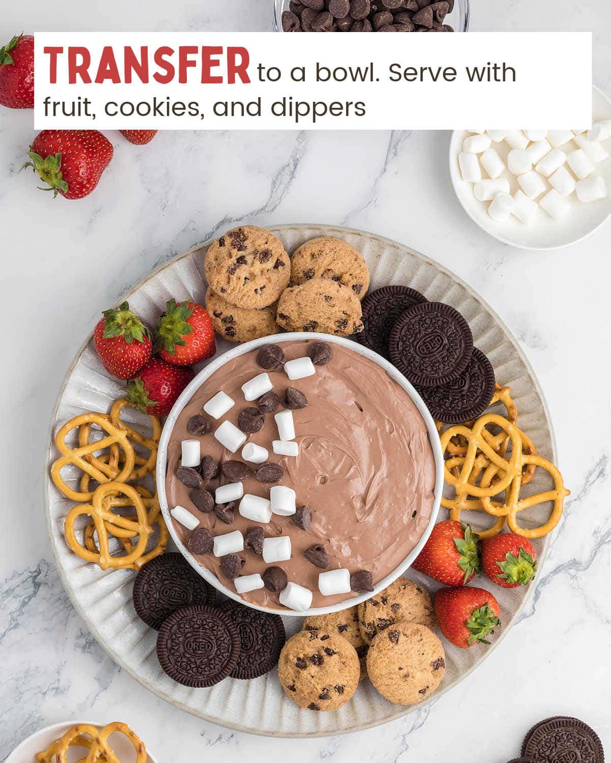 Hot chocolate dip in a bowl on a serving platter topped with mini marshmallows and chocolate tops. Around the bowl are pretzels, cookies, and strawberries.