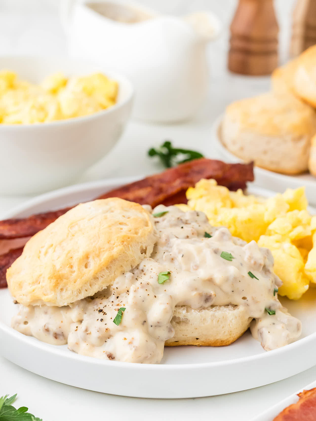 Biscuits and Gravy served with scrambled eggs and bacon on a white plate.